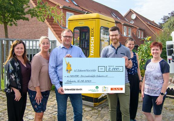 Vor der Büchertauschbox überreichten Babette Herrmann, Susanne Jenczewski und Jens Leupold vom Lions Club Fürst Niklot (v.l.) den Spendencheck an Claudia Milius, Neele Jepsen und Alexander Kieslich vom MENTOR e.V. (v.r.), Foto: maxpress