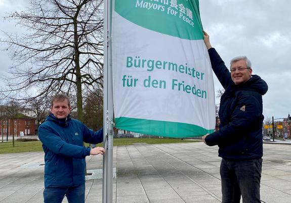 Schwerins Oberbürgermeister Rico Badenschier und der stellvertretende Stadtpräsident Daniel Meslien am Freitag auf dem Bertha-Klingberg-Platz die Flagge der Organisation „Mayors for Peace - Bürgermeister für den Frieden